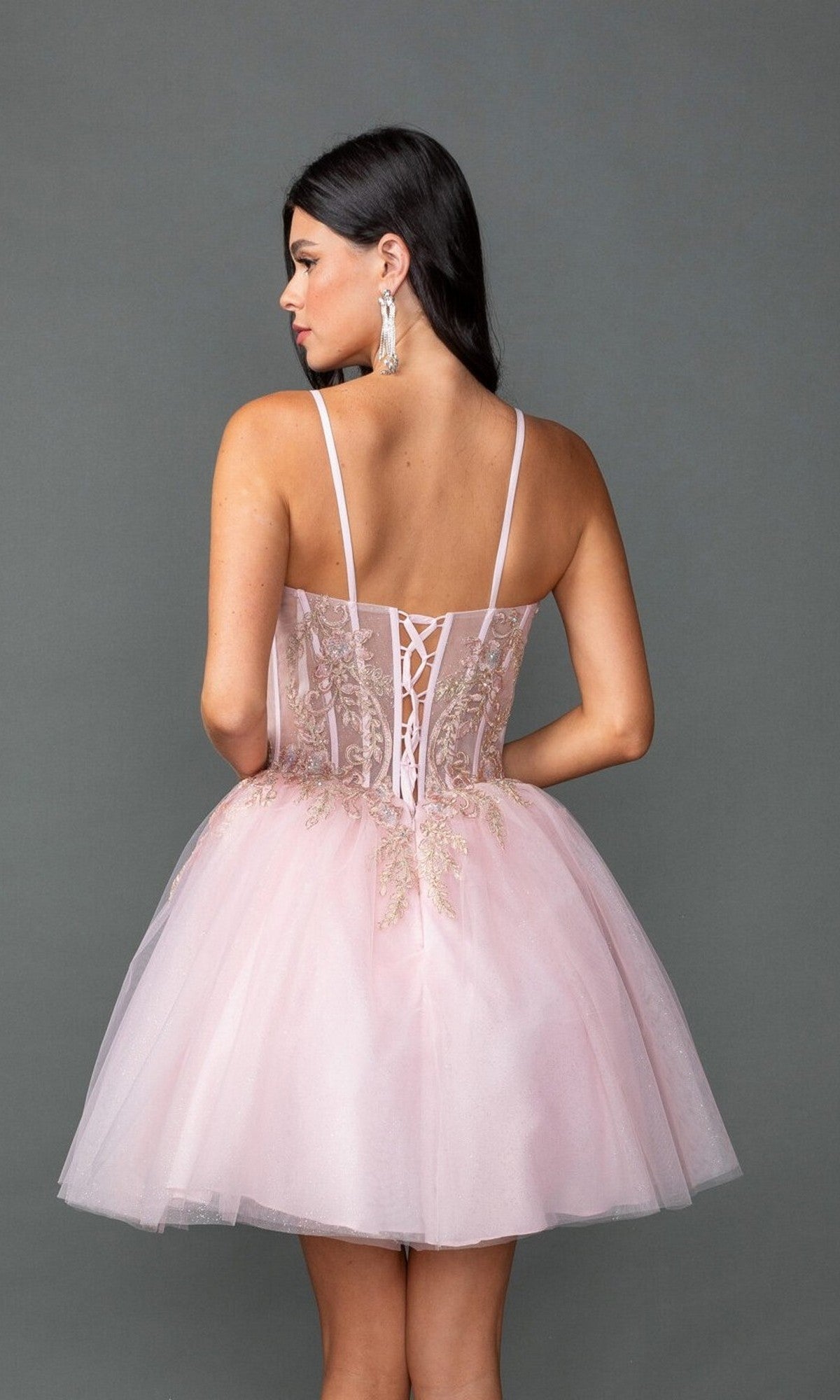 A woman faces away in the Short Prom Dress: Dancing Queen 3424, featuring a light pink hue, thin straps, and beaded lace embroidery on the bodice, set against a plain gray background.